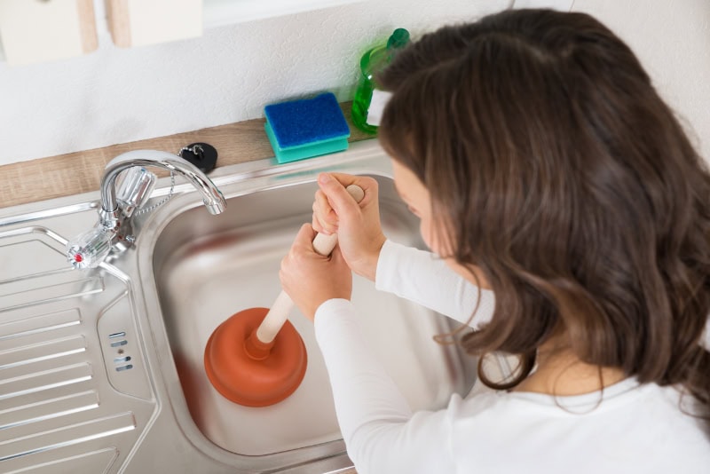 Why Do My Drains Keep Clogging? Woman Using Plunger In Kitchen Sink At Home